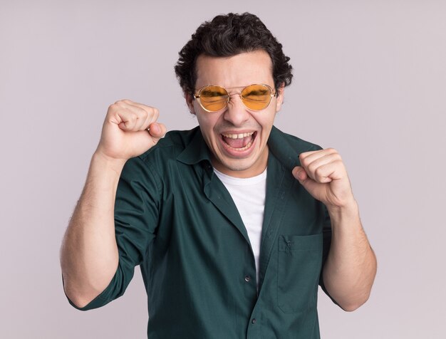 Hombre joven en camisa verde con gafas feliz y emocionado apretando los puños regocijándose de su éxito de pie sobre la pared blanca