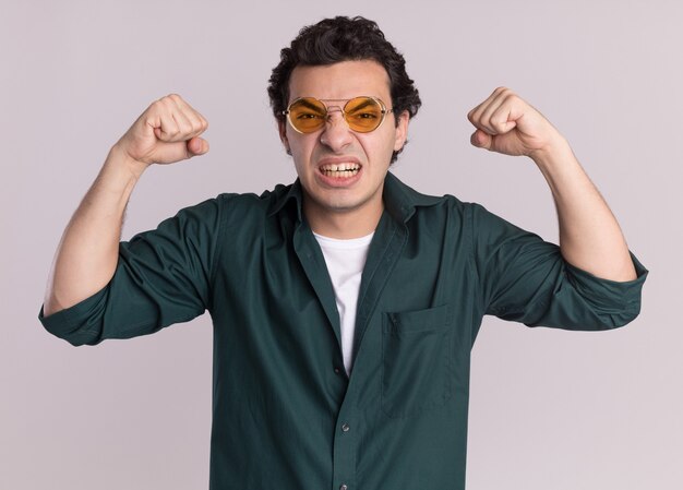 Hombre joven en camisa verde con gafas levantando puños mirando al frente con cara enojada frustrado volviéndose loco de pie sobre la pared blanca