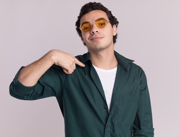 Hombre joven en camisa verde con gafas mirando al frente con expresión de confianza apuntando a sí mismo de pie sobre la pared blanca