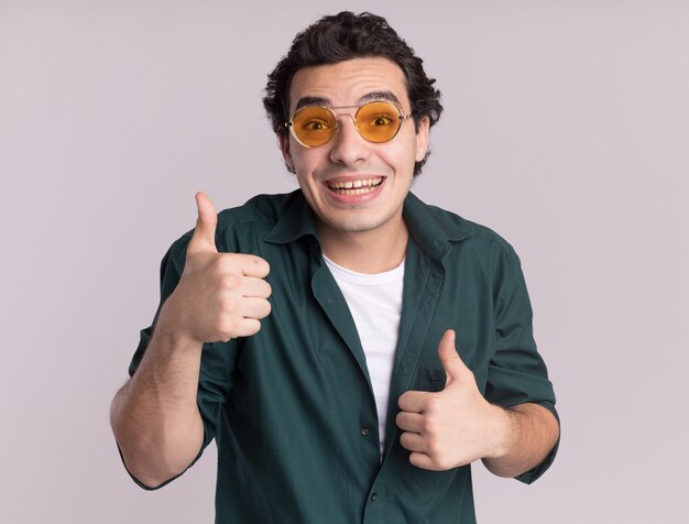 Hombre joven en camisa verde con gafas mirando al frente feliz y positivo sonriendo alegremente mostrando los pulgares para arriba de pie sobre la pared blanca