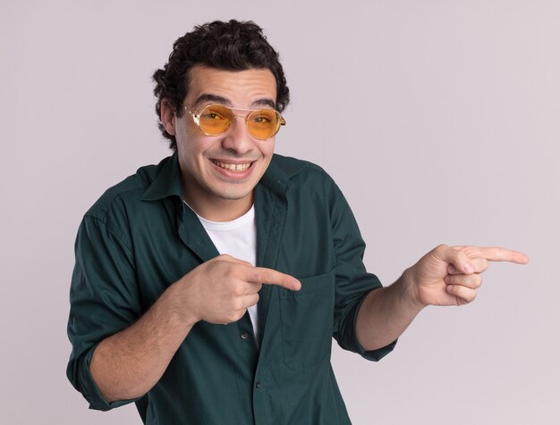 Hombre joven en camisa verde con gafas mirando al frente sonriendo astutamente señalando con los dedos índices hacia el lado de pie sobre la pared blanca