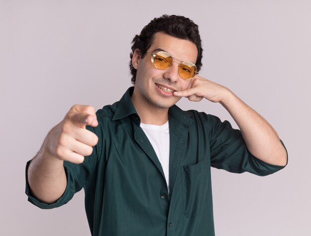 Hombre joven con camisa verde con gafas mirando al frente con una sonrisa en la cara apuntando con el dedo índice a la cámara haciendo gesto de llamarme de pie sobre la pared blanca