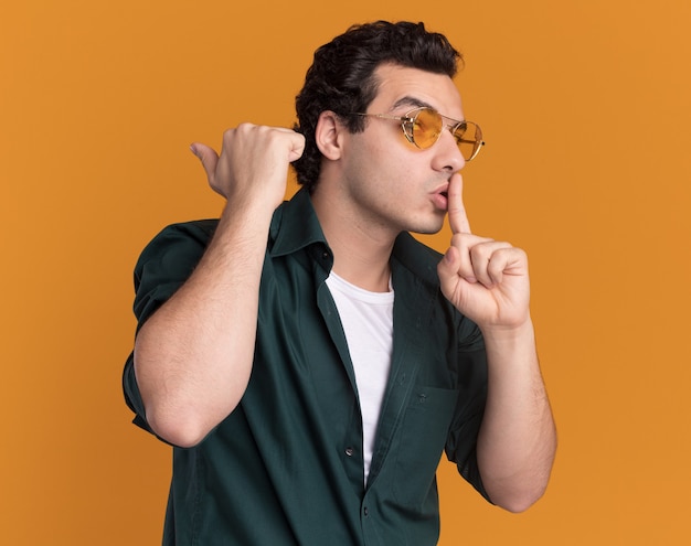 Hombre joven en camisa verde con gafas mirando a un lado haciendo gesto de silencio con el dedo en los labios de pie sobre la pared naranja
