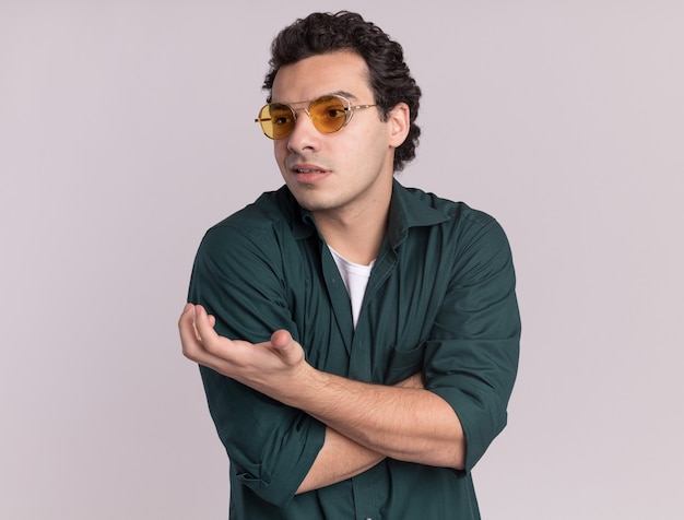 Foto gratuita hombre joven en camisa verde con gafas mirando a un lado levantando el brazo con disgusto e indignación de pie sobre la pared blanca