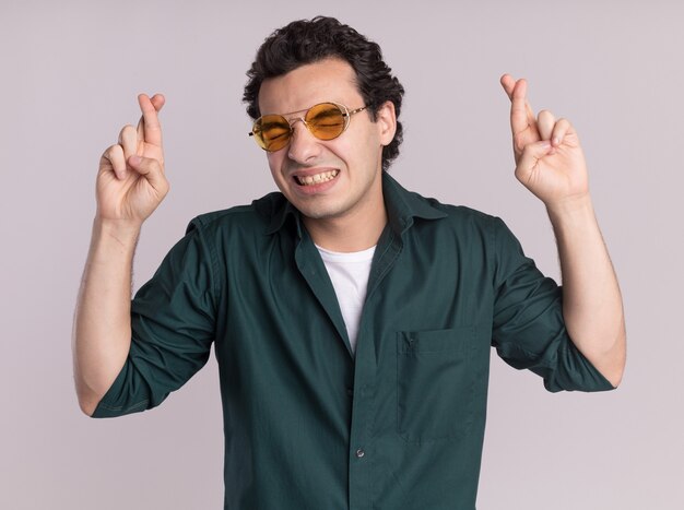 Hombre joven en camisa verde con gafas con los ojos cerrados cruzando los dedos con expresión de esperanza haciendo un deseo deseable de pie sobre la pared blanca