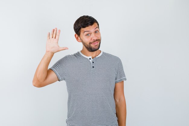 Hombre joven en camiseta agitando la mano para saludar y mirar alegre, vista frontal.