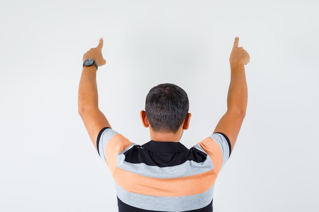 Hombre joven en camiseta apuntando hacia arriba y mirando confiado, vista posterior.