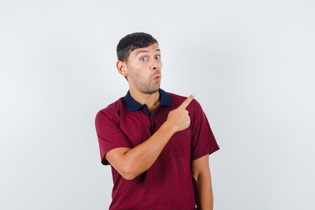 Hombre joven en camiseta apuntando hacia otro lado y mirando curioso, vista frontal.