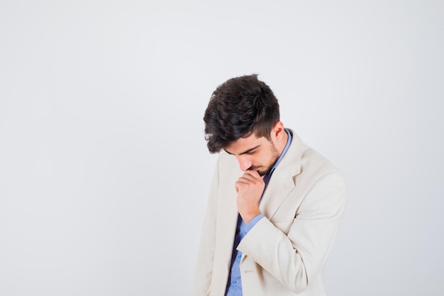 Hombre joven en camiseta azul y chaqueta de traje blanco poniendo la mano en la boca, pensando en algo y mirando pensativo