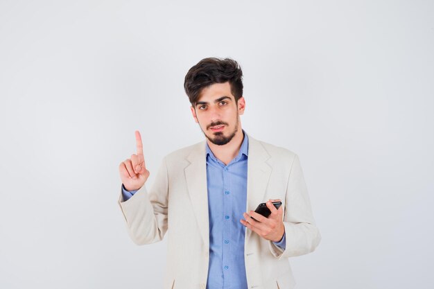 Hombre joven en camiseta azul y chaqueta de traje blanco sosteniendo teléfono inteligente y levantando el dedo índice en gesto de eureka y luciendo sensato