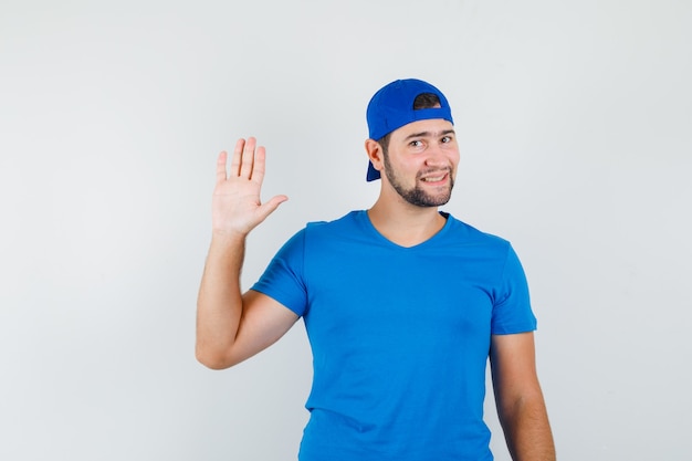 Hombre joven en camiseta azul y gorra agitando la mano para decir adiós y mirando alegre