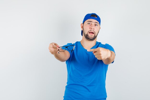 Hombre joven en camiseta azul y gorra apuntando a las gafas y mirando seguro