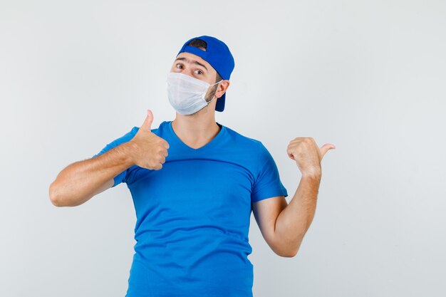 Hombre joven en camiseta azul y gorra, máscara apuntando hacia afuera con el pulgar hacia arriba y mirando confiado