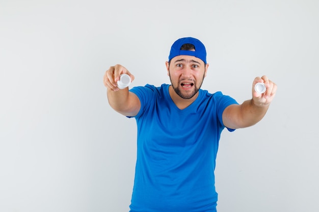 Hombre joven en camiseta azul y gorra mostrando una botella vacía de píldoras y mirando abatido.