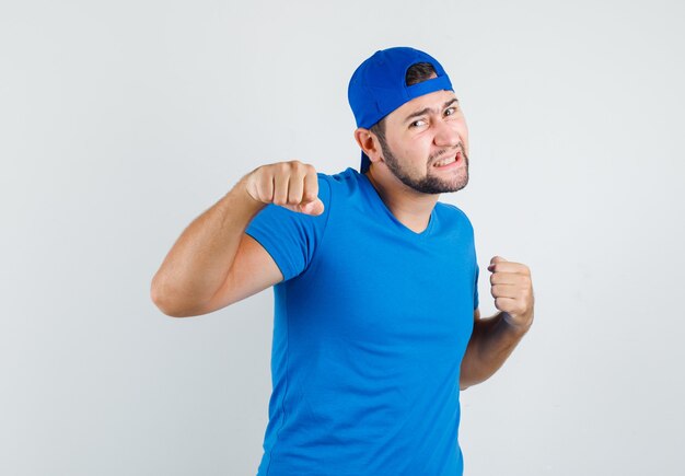 Hombre joven en camiseta azul y gorra de pie en pose de lucha y mirando pendenciero