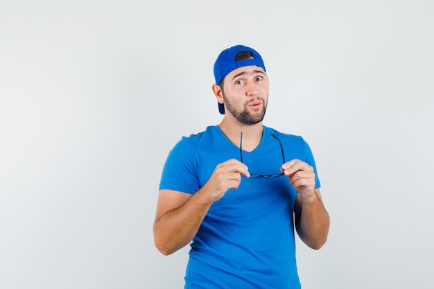 Hombre joven en camiseta azul y gorra sosteniendo gafas y mirando asombrado