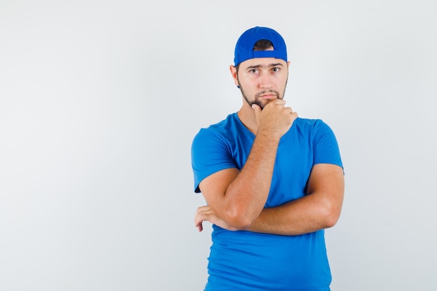 Hombre joven en camiseta azul y gorra sosteniendo su barbilla y mirando pensativo