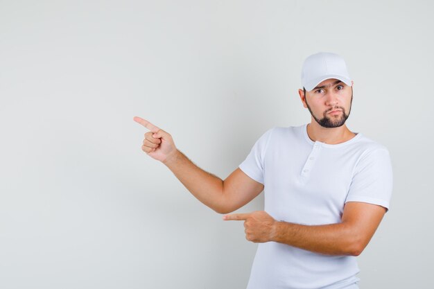 Hombre joven con camiseta blanca apuntando a un lado y mirando serio, vista frontal. espacio para texto