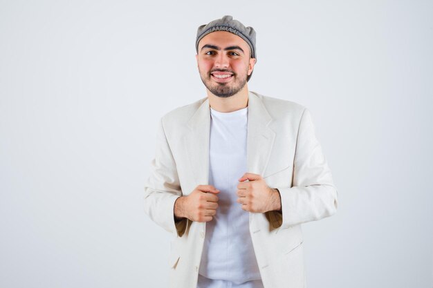 Hombre joven en camiseta blanca, chaqueta y gorra gris apretando los puños y mirando feliz