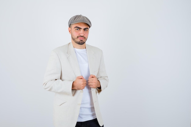 Hombre joven con camiseta blanca, chaqueta y gorra gris cogidos de la mano en la chaqueta y mirando serio