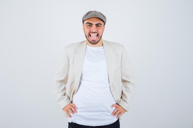 Hombre joven en camiseta blanca, chaqueta y gorra gris cogidos de la mano en la cintura, sacando la lengua y mirando feliz