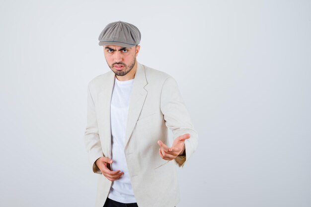 Hombre joven en camiseta blanca, chaqueta y gorra gris estirando las manos hacia el frente y mirando serio