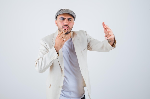 Hombre joven en camiseta blanca, chaqueta y gorra gris fumando cigarrillos y estirando la mano hacia el frente y mirando enojado
