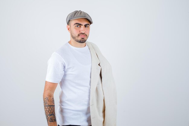 Hombre joven en camiseta blanca, chaqueta y gorra gris posando al frente con chaqueta en el hombro y mirando serio