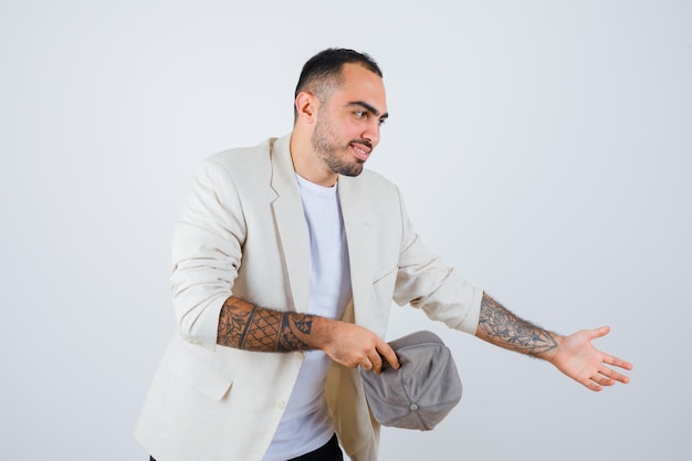 Hombre joven con camiseta blanca, chaqueta y gorra gris sosteniendo la gorra en la mano y estirando la mano como invitando a alguien y mirando feliz, vista frontal.