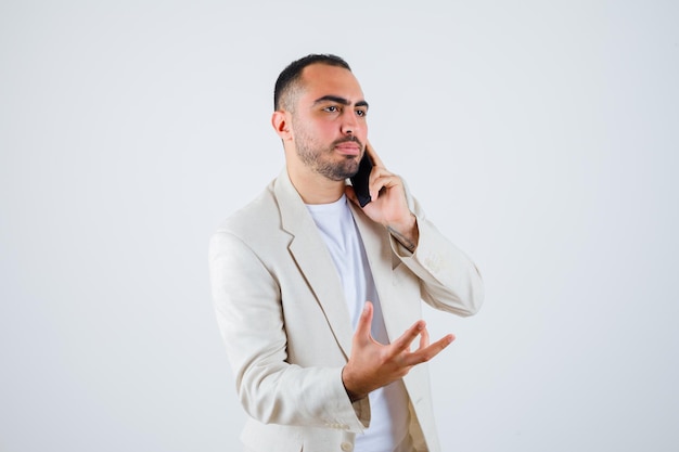 Hombre joven en camiseta blanca, chaqueta hablando por teléfono y estirando la mano como sosteniendo algo y mirando serio, vista frontal.