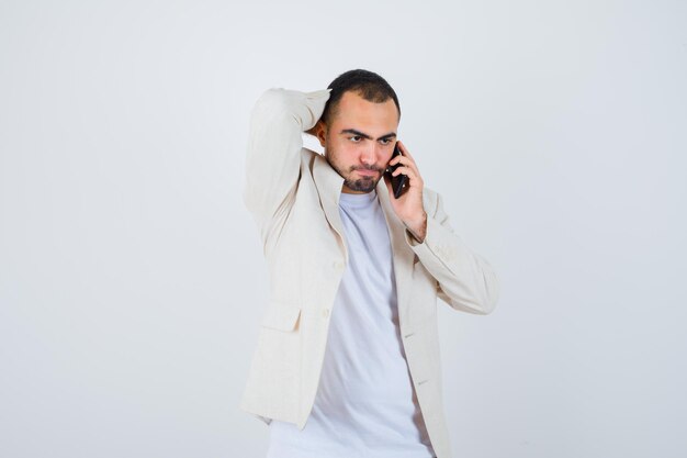 Hombre joven en camiseta blanca, chaqueta hablando por teléfono y poniendo la mano en la cabeza y mirando serio, vista frontal.