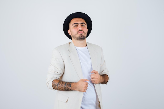 Hombre joven con camiseta blanca, chaqueta y sombrero negro cogidos de la mano en la chaqueta y mirando serio, vista frontal.