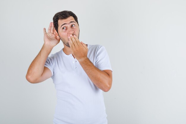 Hombre joven en camiseta blanca escuchando algo confidencial y mirando sorprendido