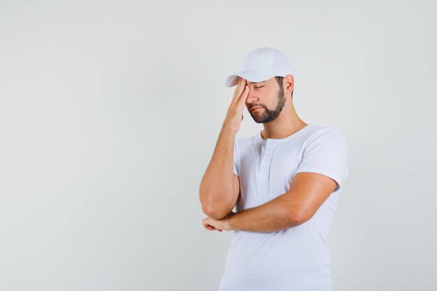 Hombre joven con camiseta blanca frotándose la frente y mirando soñoliento, vista frontal. espacio libre para tu texto