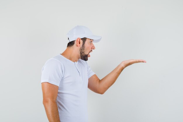 Hombre joven con camiseta blanca, gorra levantando la mano en gesto de interrogación a un lado y mirando serio.