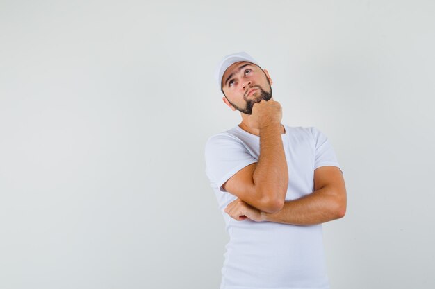 Hombre joven con camiseta blanca, gorra mirando a otro lado mientras sostiene el puño en la mandíbula y mira pensativa, vista frontal.