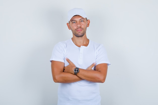 Hombre joven en camiseta blanca, gorra de pie con los brazos cruzados, vista frontal.