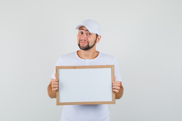 Hombre joven en camiseta blanca, gorra de pie con pizarra blanca y mirando positivo, vista frontal.