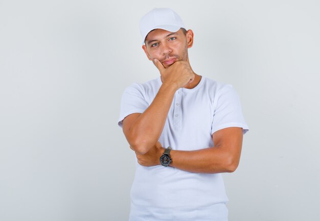 Hombre joven con camiseta blanca, gorra sosteniendo la mano en la barbilla y mirando confiado, vista frontal.