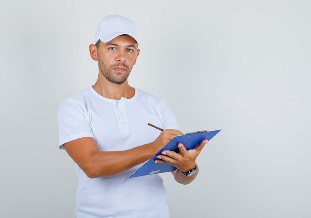 Hombre joven en camiseta blanca y gorra tomando notas en el portapapeles, vista frontal.