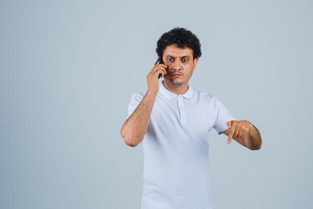 Hombre joven en camiseta blanca hablando por teléfono móvil, apuntando hacia abajo y mirando nervioso, vista frontal.