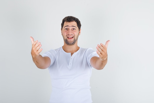 Hombre joven en camiseta blanca invitando a venir con las manos y mirando feliz