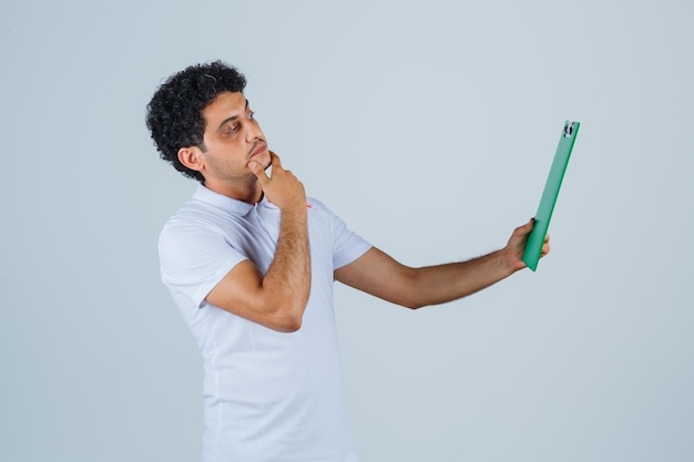 Hombre joven en camiseta blanca y jeans con cuaderno y bolígrafo, mirando el cuaderno y mirando pensativo, vista frontal.