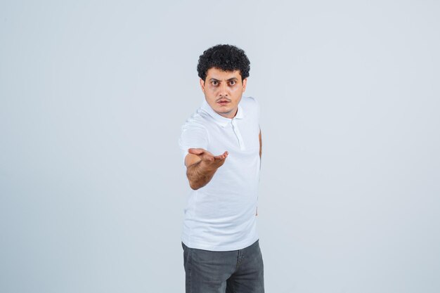 Hombre joven en camiseta blanca y jeans estirando las manos hacia la cámara y mirando serio, vista frontal.