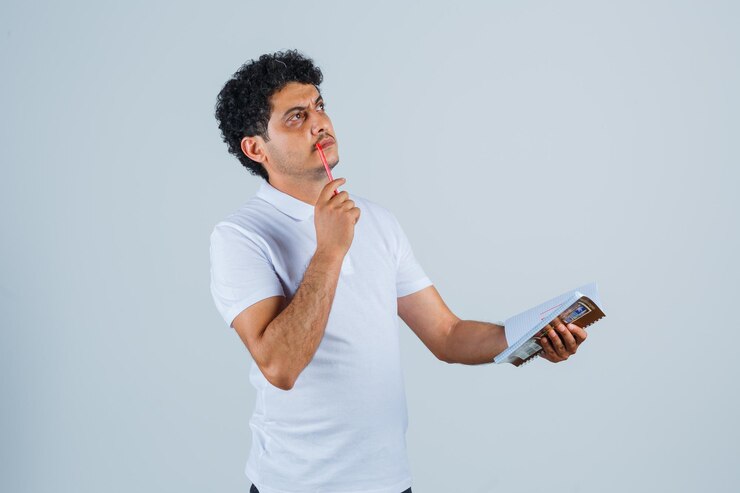 Foto gratuito hombre joven en camiseta blanca y jeans mordiendo la pluma, sosteniendo el cuaderno y pensando en algo y mirando pensativo, vista frontal.