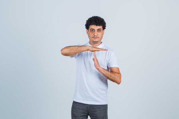 Hombre joven en camiseta blanca y jeans mostrando gesto de descanso y mirando serio, vista frontal.