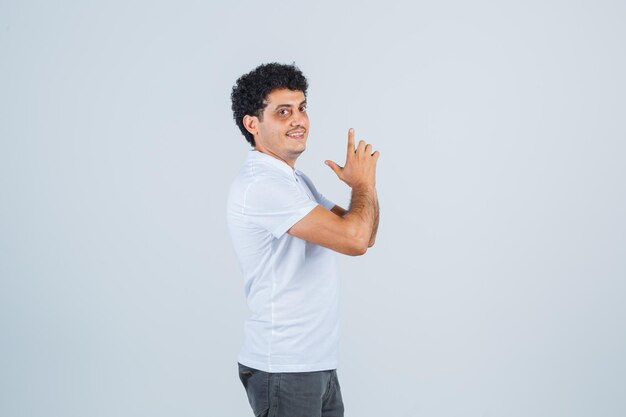 Hombre joven en camiseta blanca y jeans mostrando gesto de pistola y mirando feliz, vista frontal.