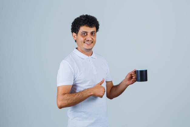 Hombre joven en camiseta blanca y jeans mostrando el pulgar hacia arriba y sosteniendo una taza de té y mirando feliz, vista frontal.