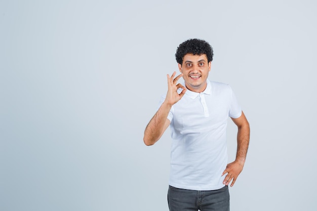 Hombre joven en camiseta blanca y jeans mostrando signo ok mientras sostiene la mano en la cintura y se ve feliz, vista frontal.