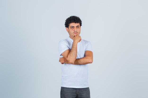 Hombre joven en camiseta blanca y jeans de pie en pose de pensamiento, con la barbilla inclinada en la mano y mirando pensativo, vista frontal.